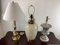 Photo of three assorted table lamps including a brass and white lamp with white shade, a white ceramic lamp with brass fittings, and a floral decorated ceramic lamp base.