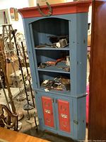 Full view of vintage painted corner cabinet with open shelves displaying miscellaneous items and lower red paneled doors with horseshoe decorations.