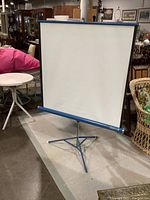 Front view of the white projector screen set up on blue metal tripod stand in vintage environment with surrounding furniture.