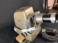 Side view of the vintage Aldis slide projector showing metal casing, lens, slide holder, and power cord.