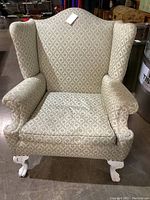 Front view of vintage wingback chair showing winged sides, rolled arms, patterned upholstery, and white painted carved wooden legs with claw-and-ball feet.