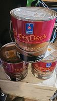 Three cans of Sherwin-Williams SuperDeck exterior solid deck stain stacked together, two large and one half gallon, with visible labels showing the brand and product type.