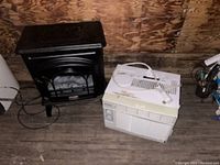 Photo showing black electric fireplace heater and white window air conditioner side by side on wooden floor.