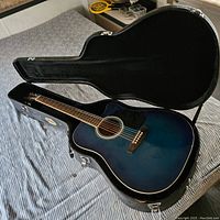 Blue acoustic guitar inside hard black case on bed, showing full guitar body and neck with strings and bridge.