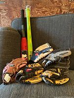 Photo showing 2 kids soft baseball bats and 5 baseball gloves arranged on a chair.
