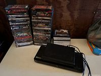 Two black DVD players with power cords and two tall stacks of DVD cases on a white surface against a wooden background.