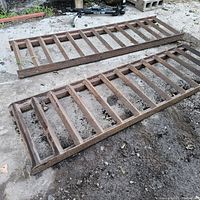 Pair of heavy metal equipment ramps laid on the ground, showing rust and weathering with a ladder-like design.