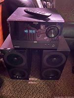 Front view of the ION home stereo unit with remote control on top and two black speakers below on carpet