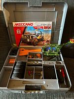 Meccano Off-Road Truck kit parts laid inside plastic carrying case including metal parts, wheels, screws, and instruction manual along with two assembled models (one green dinosaur-like and one small vehicle).