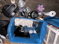 Top view of blue plastic bin filled with white kitchen towels, glassware, and some kitchen appliances and accessories on floor.