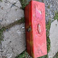 Closed red metal toolbox with handle and latch, shows wear and dirt.