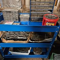 Full view of the blue rolling cart on wheels showing metal organizers, containers, and cardboard boxes with screws, nails, and pegboard hooks.