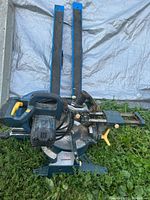 Blue and black 10 inch miter saw with yellow adjustment handles on grass with two blue folding saw horses with black padding in the background.
