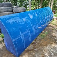 Full side view of long blue steel frame awning with shiny reflective surface. Outdoor setting with tires and trees in background.