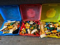 Photo of three LEGO storage bins (blue, red, yellow) filled with wooden train tracks and wooden train cars with accessories arranged inside each bin.