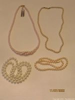 Group view of four pearl necklaces on a flat surface in soft lighting showing varying lengths, bead sizes, and colors.