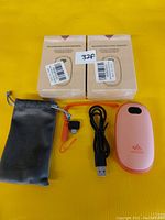 Two boxed Lingaa rechargeable handwarmers, one pink handwarmer, USB charging cable, wrist strap with orange hook, and gray carrying sleeve arranged on a yellow cloth background.