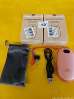 Two boxed rechargeable handwarmers, a light pink handwarmer out of the box, black micro USB charging cable, orange wrist strap, and a gray drawstring carrying pouch on a yellow surface.