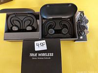 Photo of two sets of true wireless stereo sport earbuds in black with their charging cases open and USB cables beside them, displayed on a yellow surface.