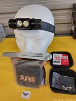 Slonik headlamp mounted on a white mannequin head displaying the adjustable strap and dual LED lights, with accompanying carrying case, battery, charger, and sizing clips arranged neatly on a yellow surface.