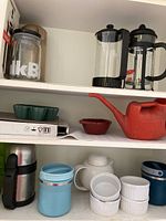Shelf with two Bodum French press coffee makers, green leaf-shaped dishes, red plastic watering can, tiny red bowl, and silver and blue insulated containers.