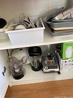Shelf with Cuisinart blender, coffee grinder, Omega juicer, and a box with kitchen gadgets. Stack of baking dishes above.