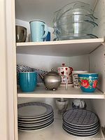 Shelf displaying assorted mugs, Mikasa Avery Medley patterned stoneware plates and bowls, marble mortar and pestle, and small teapots.