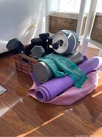 Photo shows five hex dumbbells in a woven basket along with an AB Carver wheel, two mats rolled (purple and pink), a black foam roller, green resistance band, and a small blue exercise ball.