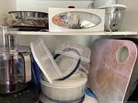 Shelf showing Hamilton Beach food processor with attachments and assorted plastic storage containers with lids, pink cutting board