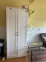 Full view of tall white kitchen storage cabinet with two doors, standing next to a wicker chair and glass-top metal shelf unit.