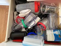 Drawer filled with various plastic measuring and blender cups with colored lids, blender base, and small kitchen containers.