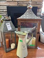 Photo showing two lanterns and a cream-colored flower vase jug placed on floor, candles inside lanterns visible