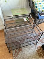 Three dark metal mesh stands stacked together on a floor rug near a chair, showing wire mesh shelves and black metal frame.