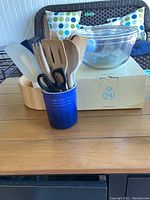 Wide view showing blue ceramic crock holding wooden and plastic kitchen utensils, wooden salad bowl, glass Pyrex mixing bowls stacked on box