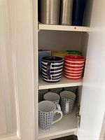 Cabinet shelf displaying four colorful M and S striped mugs (navy blue, red, yellow, green) and below two white and blue pottery mugs.