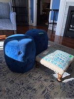 Side view of two blue tufted round ottomans and one patterned foot stool on a carpeted floor.
