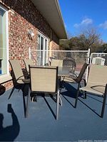 Patio set featuring rectangular glass top table with six matching metal framed chairs on a deck outside a brick house.