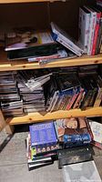 Photo showing stacks of CDs, DVDs and Blu-rays on wooden shelving and floor, varied genres and titles visible.