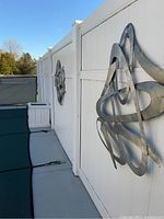 View of two matching abstract metal wall sculptures mounted on a white fence, with a white wooden planter visible in the distance.