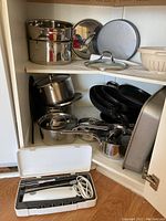 Cookware items arranged inside kitchen cabinet including stainless steel pots with lids, non-stick pans stacked, cake pans, splash screen, and ceramic colander bowl.