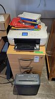 Photo showing white Canon Pixma multifunction printer on a small wooden cabinet with multiple drawers. The printer has printer papers and manuals stacked on top. Below the cabinet is a black Black & Decker paper shredder. Wall with exposed wiring in background.