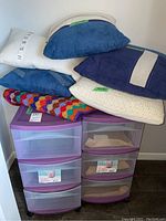 Full view of two plastic storage containers with five assorted throw pillows and a multicolored knotted throw on top.