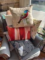 Six assorted decorative throw pillows on a chair including one with embroidered bird and various abstract patterns in red, blue, and neutral colors.