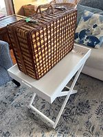 Photo showing the brown and beige woven wicker picnic basket on a white collapsible TV tray over a patterned carpet in a living area.