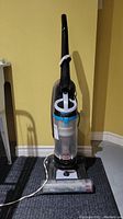 Front view of Bissell Cleanview upright vacuum showing handle, transparent dust bin with blue accents, hose attached to the side, and power cord laying on the floor.