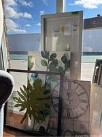 Overall view of four framed decorative wall art pieces including leaf, floral, clock with 'Home' wording, and watercolor artworks placed against a window showing outdoor background.