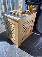 Wooden clothes hamper with lattice side panels and top, placed near a window with sunlight casting shadows. Teapot and cereal box are on top for scale. Dimensions visible on a label.