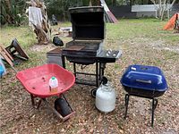 Full view of the Weber propane grill, small charcoal grill, red wheelbarrow with a jug inside, and propane tank outdoors on dirt ground.