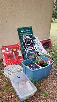 Overall photo showing assorted outdoor Christmas decorations including boxed spiral tree, Santa frame, Happy Holidays sign, poinsettia, and plastic containers with lights and accessories arranged outside against a wall.