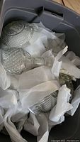Clear cut glass liquor decanter and other wrapped glassware items in protective white paper inside a plastic container